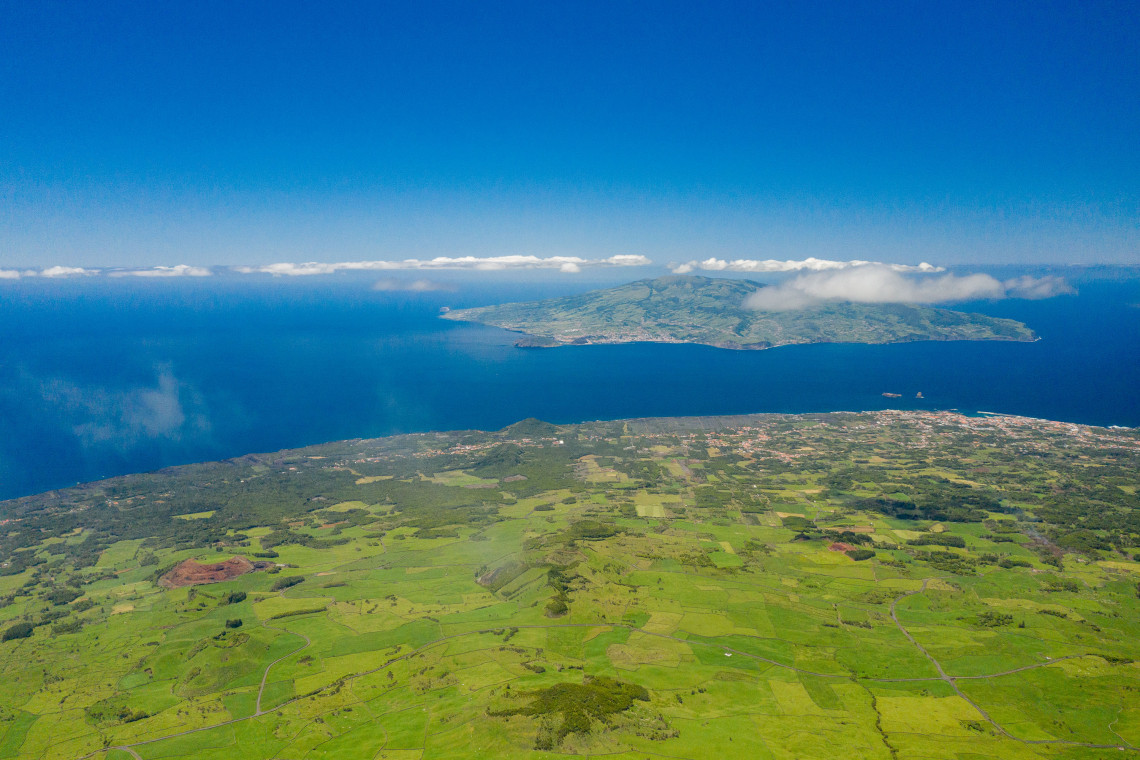 Aerial view across the channel between Pico and Faial islands, showing Pico&rsquo;s patchwork green fields in the foreground and Faial Island surrounded by the Atlantic Ocean.